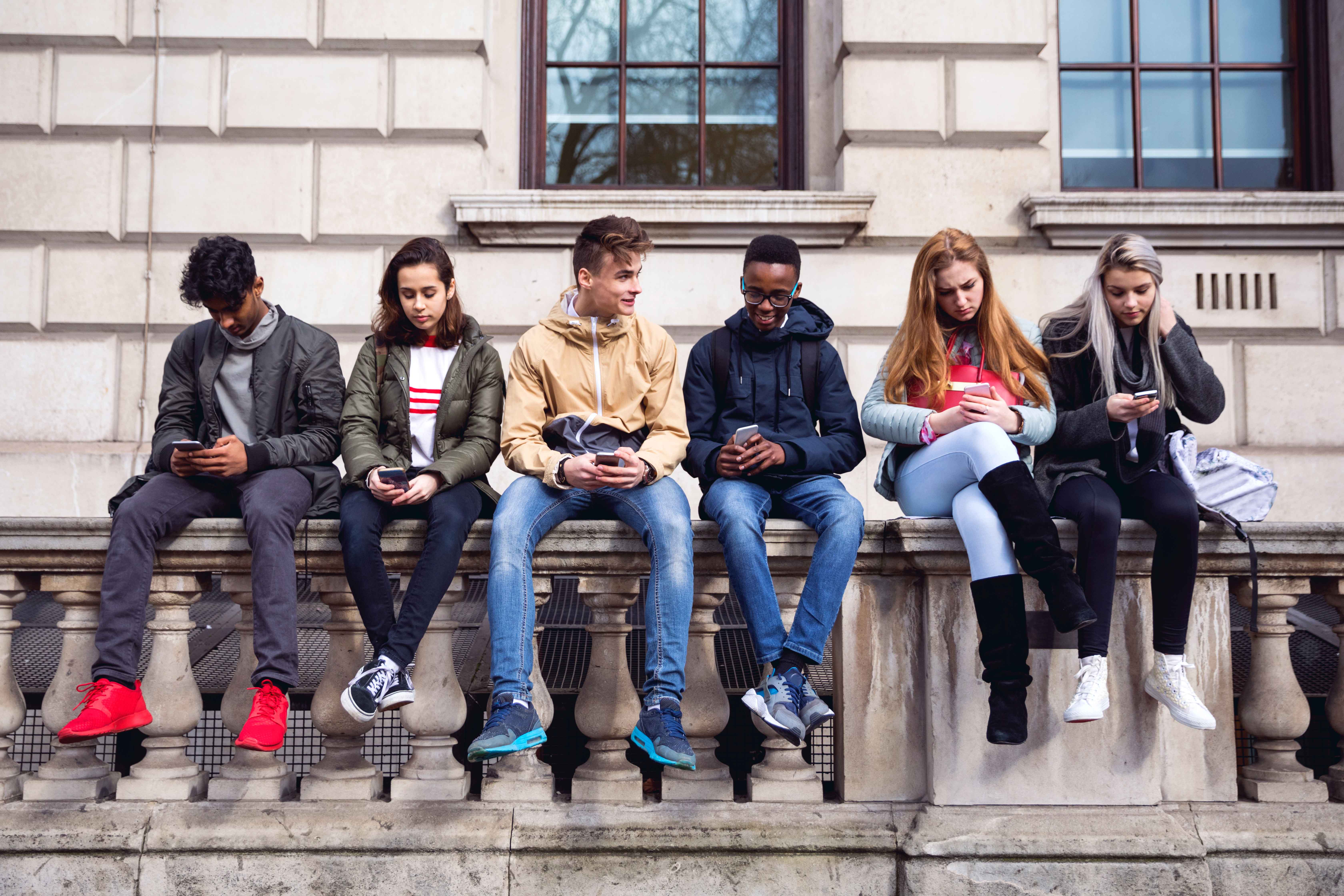 high-school-aged kids sitting along a building's stone rail, looking at mobile devices